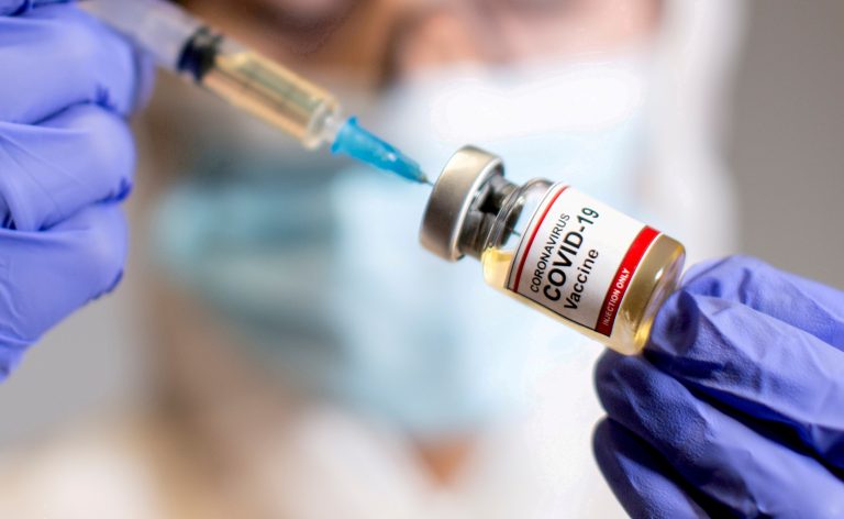 A woman holds a medical syringe and a small bottle labelled "Coronavirus COVID-19 Vaccine